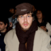 Jack Harlow wearing glasses, a brown hat, and a scarf sits in a crowd. Others around him have various styles, including tattoos and sunglasses.