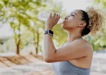 Photo of a woman drinking water from a plastic bottle in a green park surround by trees