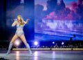Taylor Swift performs on stage in a shimmering bodysuit and boots, holding a microphone, with a large screen displaying a blue and purple sky behind her.