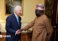 King Charles, dressed in a navy pin stripe suit, white shirt and sky blue tie, shakes hands with President Tinubu, who wears a light brown outfit and colourful kufi. They stand in a brightly lit room, with pale yellow walls adorned with gold-framed paintings and wooden floors decorated with a dark red rug.