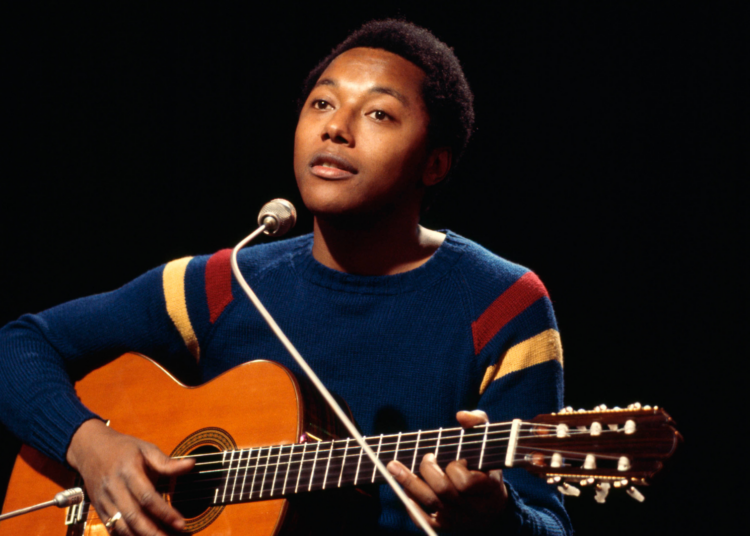 Labi Siffre playing an acoustic guitar and singing into a microphone, wearing a colorful sweater, set against a dark background.