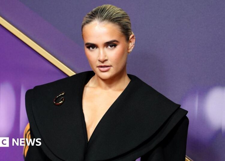 Moly-Mae Hague on a red carpet. Background is purple and gold and the social media star wears a black wide cut collared dress with her blonde hair scraped back.