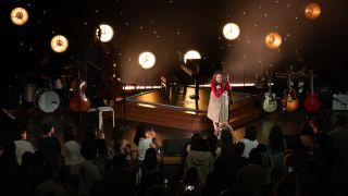 A woman sings on a stage containing a number of guitars, a drum set and other instruments, in front of a lightscape of moons and stars.