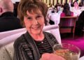 An elderly woman with short hair is seated in a dining area, holding a glass in her hand. In the background, there are several p