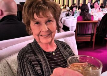 An elderly woman with short hair is seated in a dining area, holding a glass in her hand. In the background, there are several p