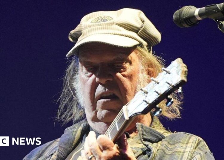 Neil Young is playing a guitar on stage while wearing a green cap and checkered jacket. He has long hair and is standing next to a microphone.