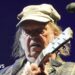 Neil Young is playing a guitar on stage while wearing a green cap and checkered jacket. He has long hair and is standing next to a microphone.