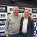 An old man and a younger man stand together in front of 2GB signage in an office