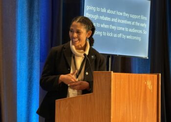 Woman with light brown skin stands, smiling, behind a wooden lectern. A screen behind her features text about film incentives. A blue curtain hangs in background.