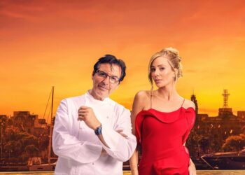 An adult male and an adult female are standing side by side against the backdrop of a cityscape, with the setting sun casting a warm glow across the scene.