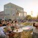 People sitting at tables and chairs on lawn with large building in background