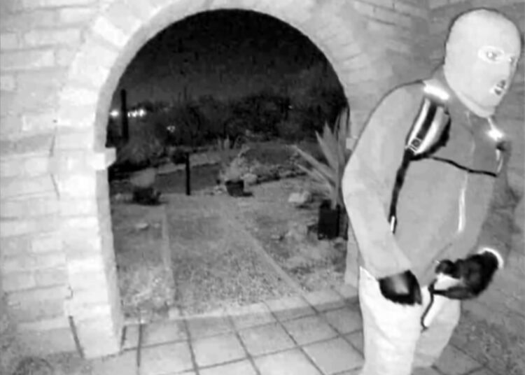 Surveillance camera footage of a person wearing a ski mask, gloves, and a backpack, standing on a doormat in front of a brick archway.