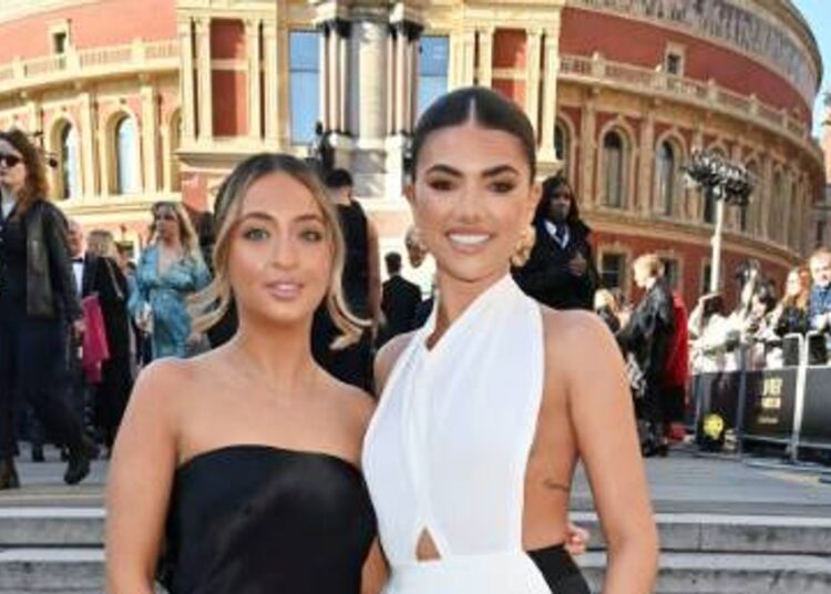 Two women are standing side by side on the steps in front of a large architectural building, possibly attending an event. The woman on the left is wearing a black dress, while the woman on the right is wearing a white dress. Several people are walking behind them, some of whom are wearing sunglasses.