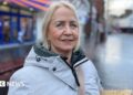 A woman with light hair looking at the camera, she is wearing a grey coat which is zipped up to the neck. She is standing on a high street with shops in the background.