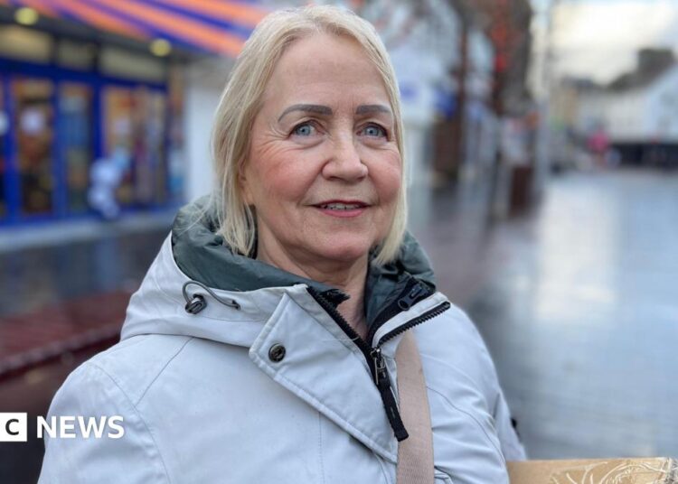A woman with light hair looking at the camera, she is wearing a grey coat which is zipped up to the neck. She is standing on a high street with shops in the background.