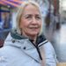 A woman with light hair looking at the camera, she is wearing a grey coat which is zipped up to the neck. She is standing on a high street with shops in the background.