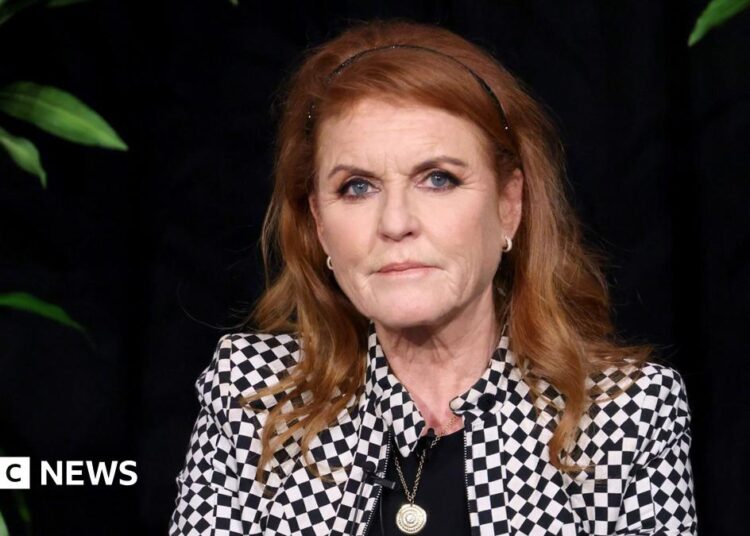 Sarah Ferguson wears a black and white checked jacket with a black top underneath, and is sitting against a black background - plants can just be seen at the side. Her red hair is pushed back with a thin black headband and she has a neutral expression on her face