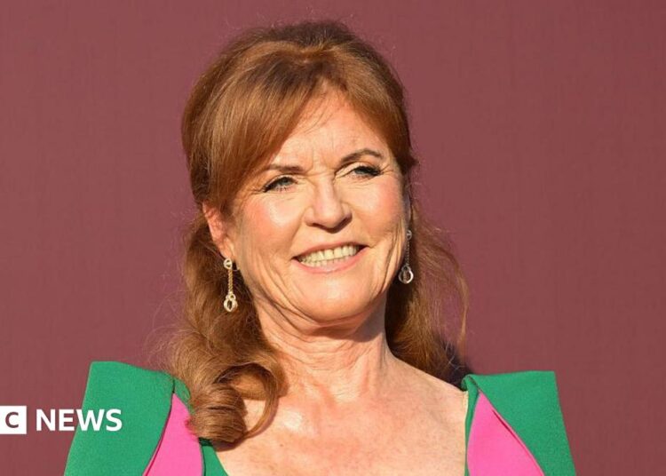 Sarah Ferguson stood in front of a dark red back drop. She is wearing a green dress with pink parts to it.