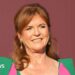 Sarah Ferguson stood in front of a dark red back drop. She is wearing a green dress with pink parts to it.