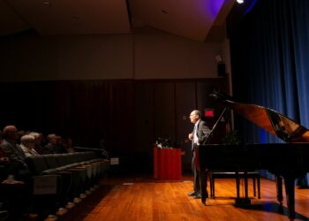 Speaker on stage beside a grand piano, addressing an audience in an auditorium.