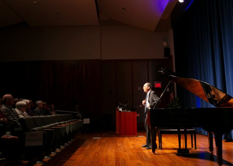 Speaker on stage beside a grand piano, addressing an audience in an auditorium.