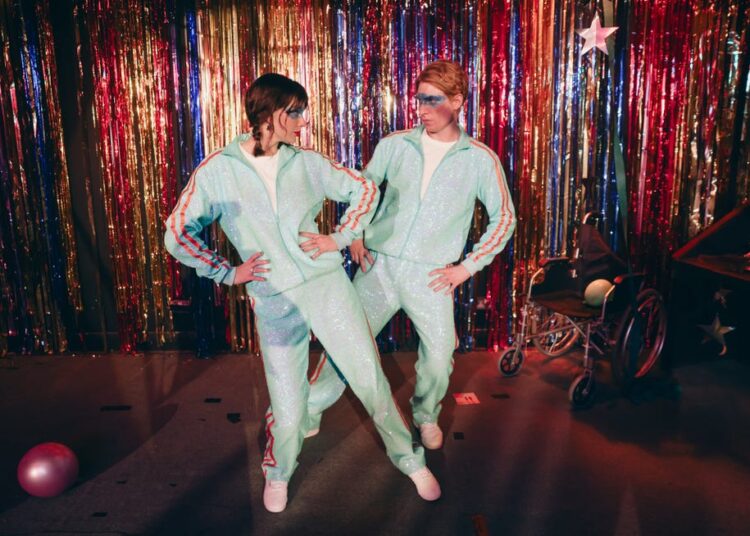 two individuals wearing matching sparkly tracksuits posing against a colorful backdrop