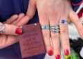 A hand holding a wooden plaque with an engraved quote and several rings.