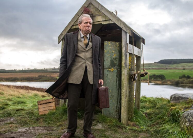 Timothy Spall standing outside a run-down bus shelter in The Last Bus