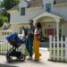 A man and a woman pushing a baby stroller outside a home with a white picket fence.