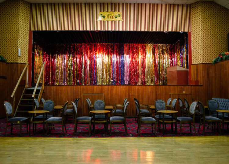 chairs and tables sat out in a hall in front of a stage with milticoloured streamers as the curtains