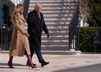 An individual dressed in a formal black coat accompanied by another person wearing a beige coat and red boots is walking in fron