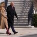 An individual dressed in a formal black coat accompanied by another person wearing a beige coat and red boots is walking in fron