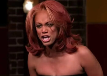 Tyra Banks on a reality show set, appearing intense and expressive, with voluminous styled hair and a strapless dress