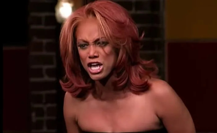 Tyra Banks on a reality show set, appearing intense and expressive, with voluminous styled hair and a strapless dress
