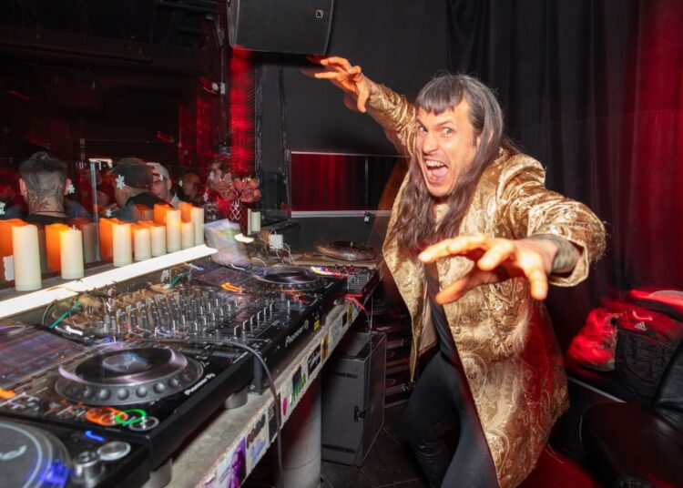 Photo of a man next to CDjs wearing a golden coat.
