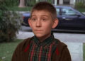 Young boy with a curious expression stands outdoors wearing a plaid shirt and a jacket, likely in a suburban setting