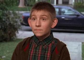 Young boy with a curious expression stands outdoors wearing a plaid shirt and a jacket, likely in a suburban setting