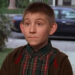 Young boy with a curious expression stands outdoors wearing a plaid shirt and a jacket, likely in a suburban setting