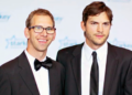 Two men in black suits stand together at an event. One wears glasses and a bow tie, smiling, while the other wears a necktie and has a neutral expression. The background features a blue and white logo with stars.