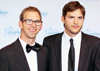 Two men in black suits stand together at an event. One wears glasses and a bow tie, smiling, while the other wears a necktie and has a neutral expression. The background features a blue and white logo with stars.