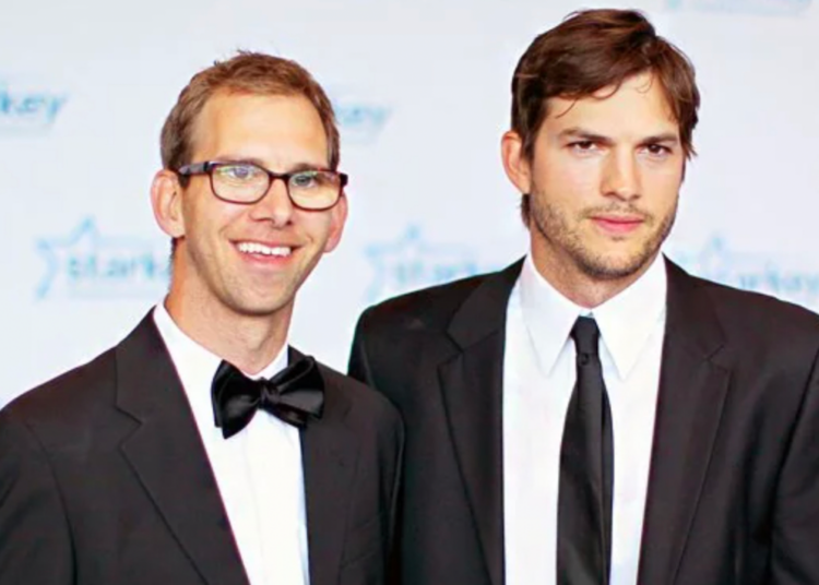 Two men in black suits stand together at an event. One wears glasses and a bow tie, smiling, while the other wears a necktie and has a neutral expression. The background features a blue and white logo with stars.