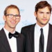 Two men in black suits stand together at an event. One wears glasses and a bow tie, smiling, while the other wears a necktie and has a neutral expression. The background features a blue and white logo with stars.