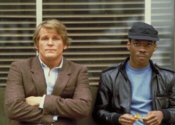 Two men stand side by side. The man on the left wears a brown blazer and looks at the camera with his arms crossed, while the man on the right wears a black leather jacket and cap, holding food in his hands. They stand in front of a window with blinds.