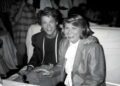 A young man and an older woman sit closely together, smiling at a table in a crowded, dimly lit room. The man has his arm around the woman, and both are dressed in formal attire.