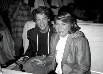 A young man and an older woman sit closely together, smiling at a table in a crowded, dimly lit room. The man has his arm around the woman, and both are dressed in formal attire.