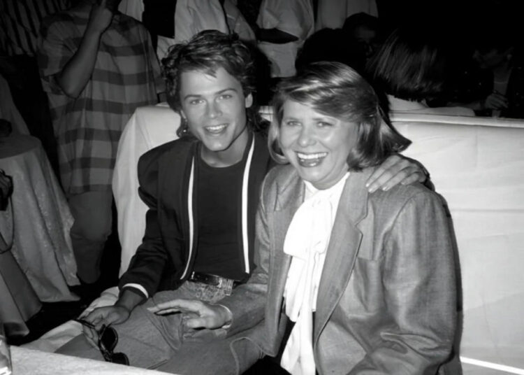 A young man and an older woman sit closely together, smiling at a table in a crowded, dimly lit room. The man has his arm around the woman, and both are dressed in formal attire.