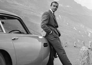 A man in a suit leans casually against a classic car parked on a mountain road, with a scenic view of mountains and valleys in the background. The image is in black and white.