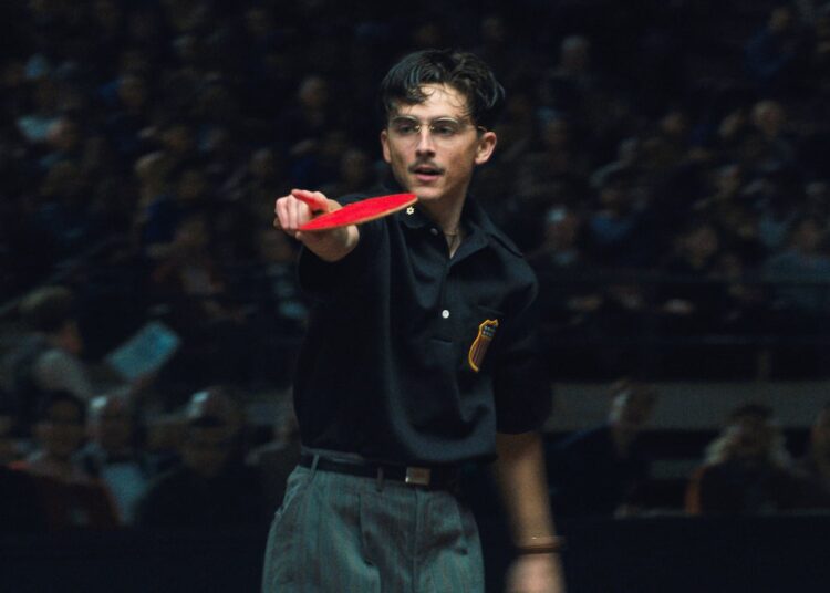 A man in a dark polo plays ping pong in front of a crowd.