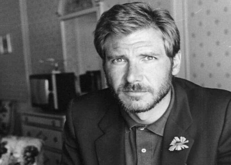 A man with short hair and a beard, wearing a suit jacket and collared shirt with a flower on the lapel, gazes seriously at the camera in a vintage-style, softly lit room.