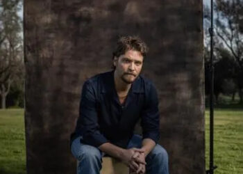 A bearded man in a blue shirt and jeans sits with his hands folded together.
